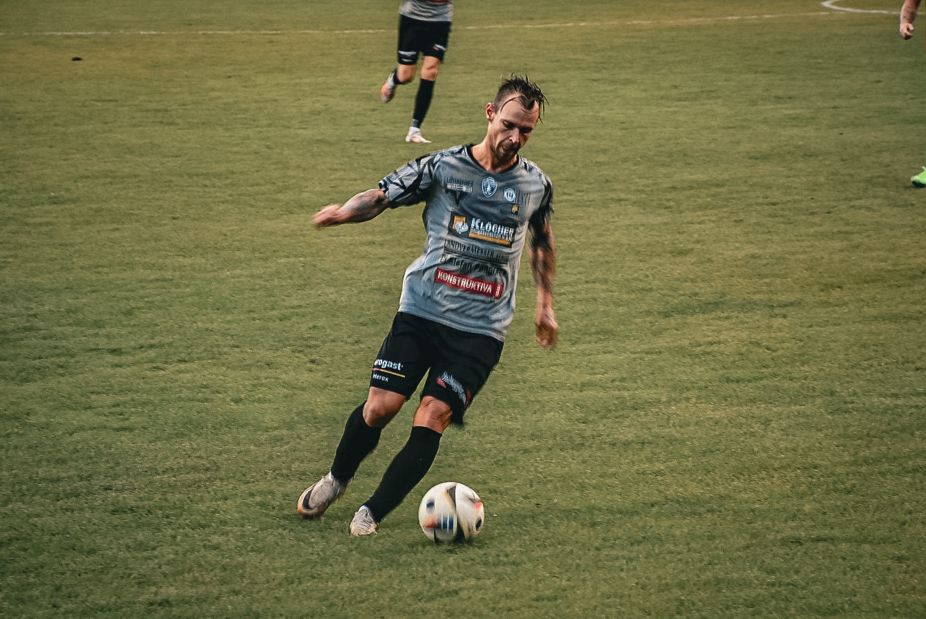 Fußballspieler in grauem Trikot dribbelt den Ball auf dem Spielfeld während eines spannenden Spiels.