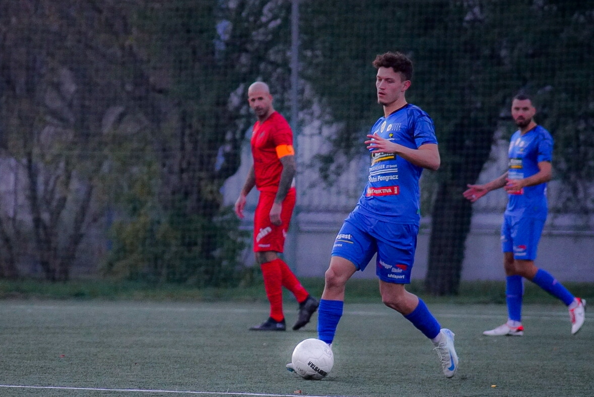 Fußballspiel auf Rasen mit Spielern in blauen und roten Trikots, Spieler führt den Ball, andere im Hintergrund.