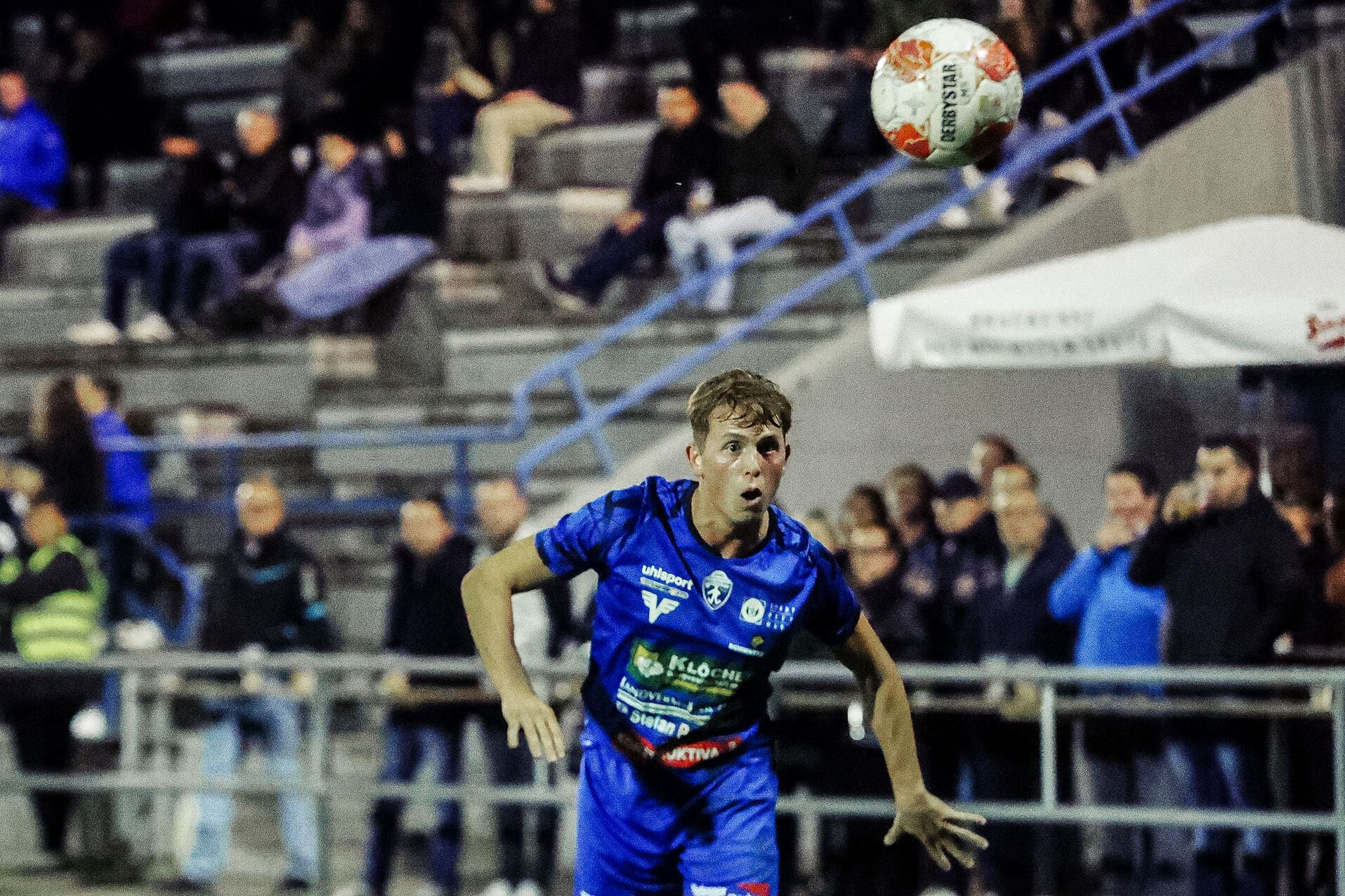 Fußballspieler im blauen Trikot konzentriert sich auf den Ball, während Zuschauer im Hintergrund sitzen.