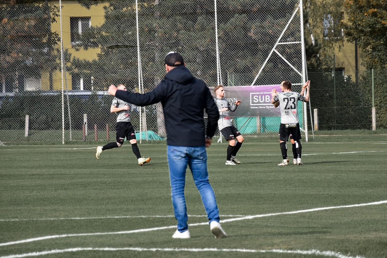 Fußballspieler jubeln auf dem Spielfeld, während der Trainer am Rand motivierend anfeuert, sonniges Wetter.