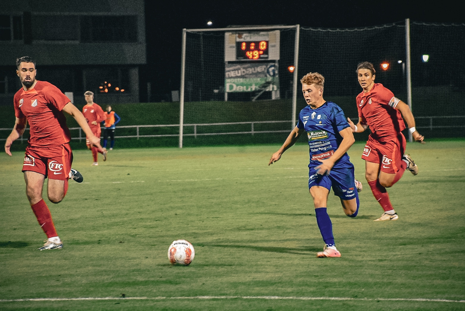 Fußballspiel bei Nacht: Spieler in Rot und Blau kämpfen dynamisch um den Ball auf dem Feld.