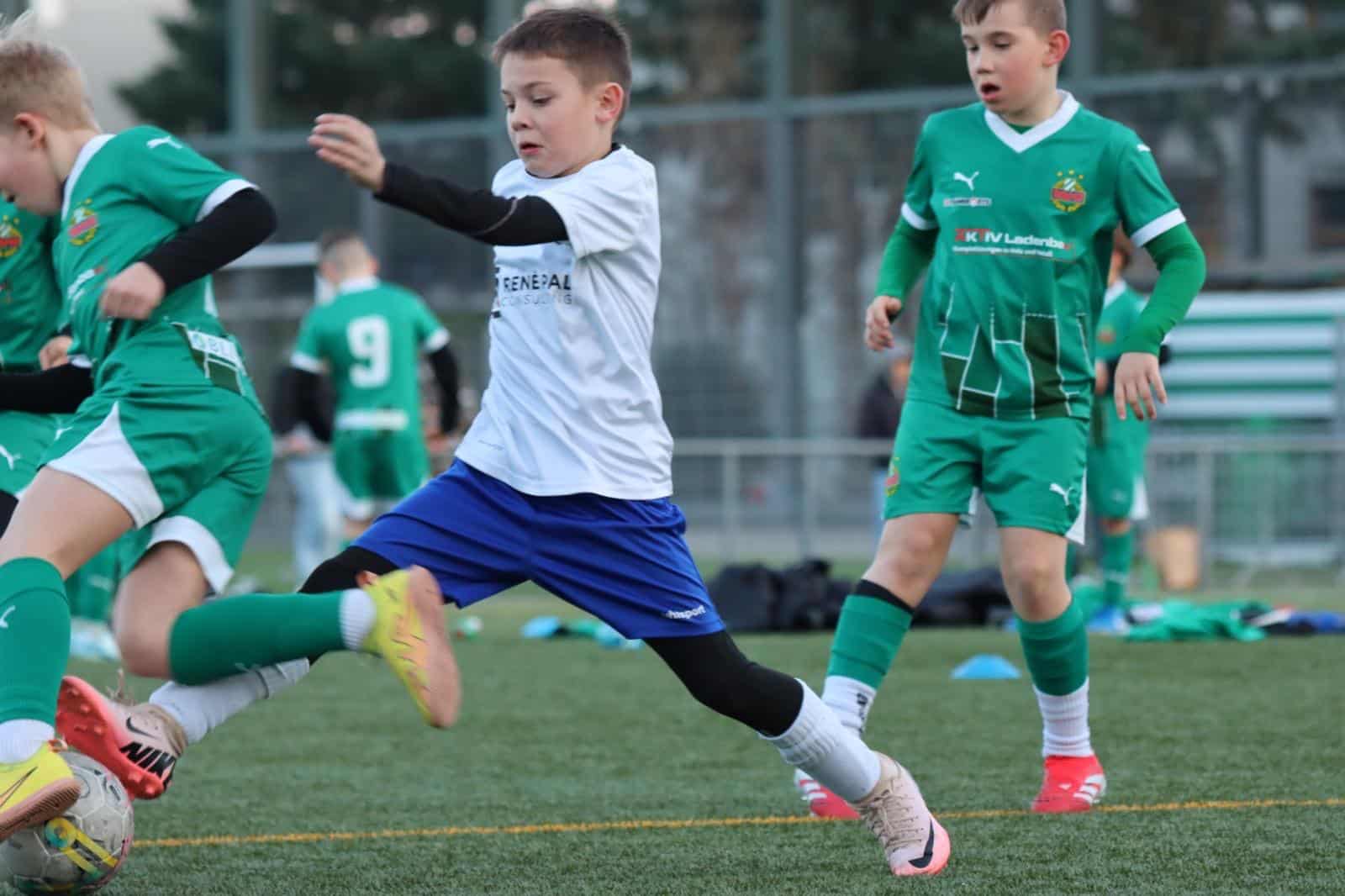 Kinder spielen intensiv Fußball auf einem Sportplatz, grüne und weiße Trikots, Action im Spiel.