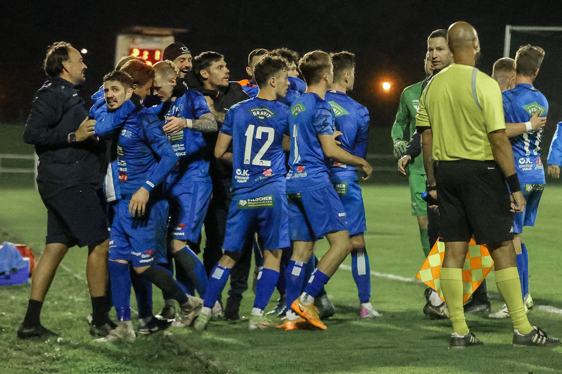 Fußballteam in blauen Trikots feiert auf dem Spielfeld nachts einen Sieg mit einem Schiedsrichter im gelben Trikot.