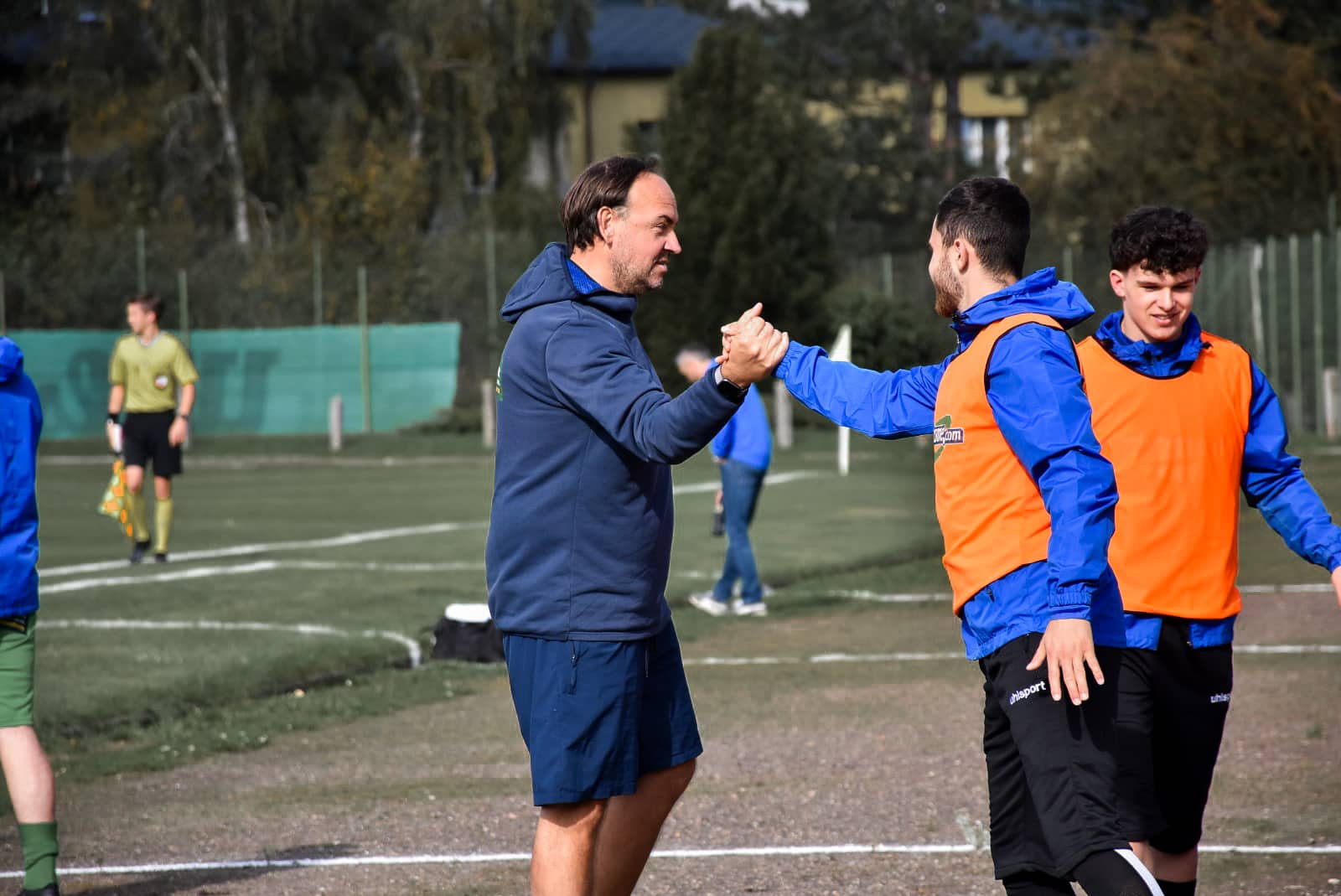 Trainer gratuliert Spieler beim Fußballspiel auf dem Feld, Team in blauen Trainingsanzügen, Emotionen des Erfolgs.