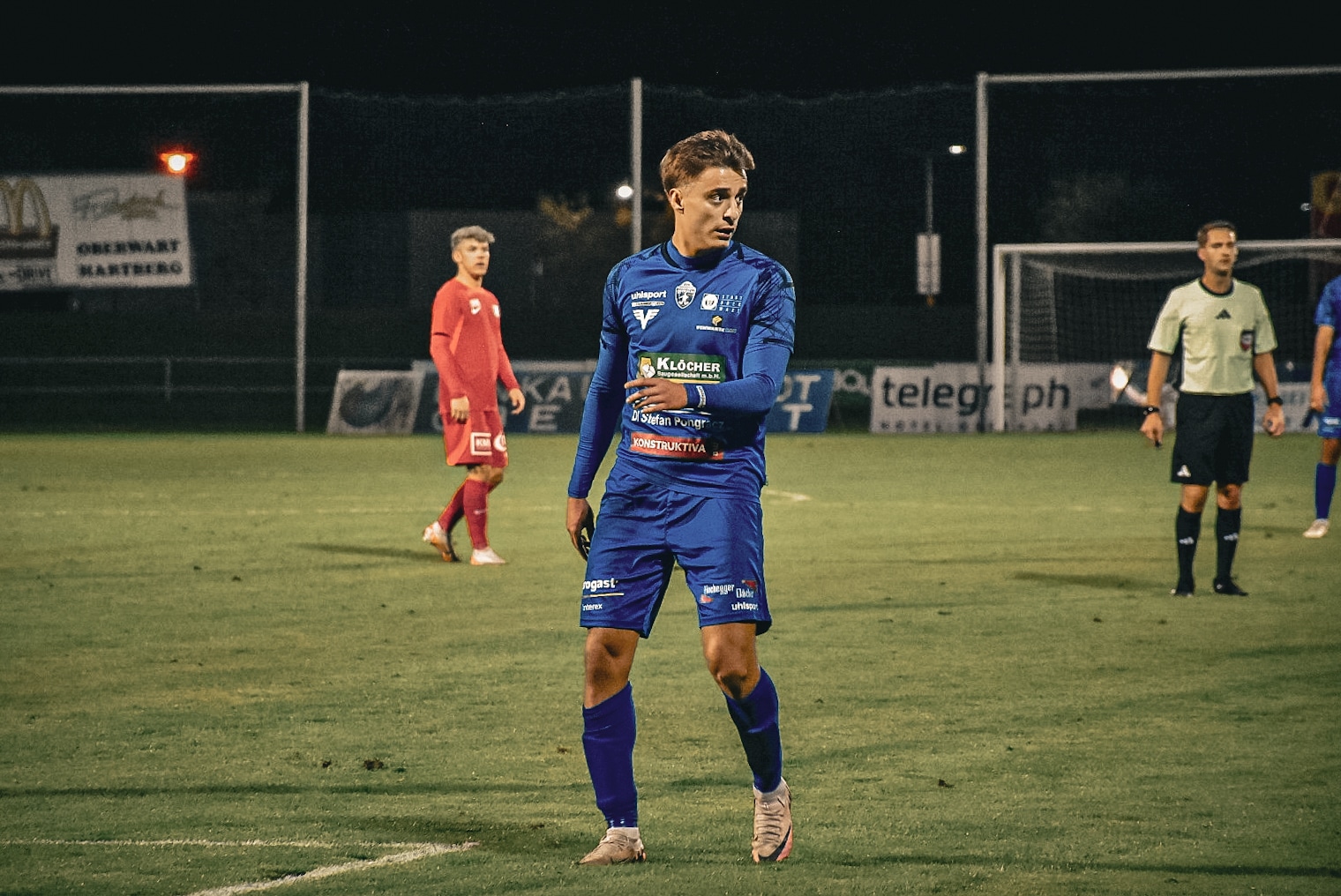 Fußballspieler in blauem Trikot auf dem Spielfeld bei Nachtspiel, neben Spielern und Schiedsrichter.