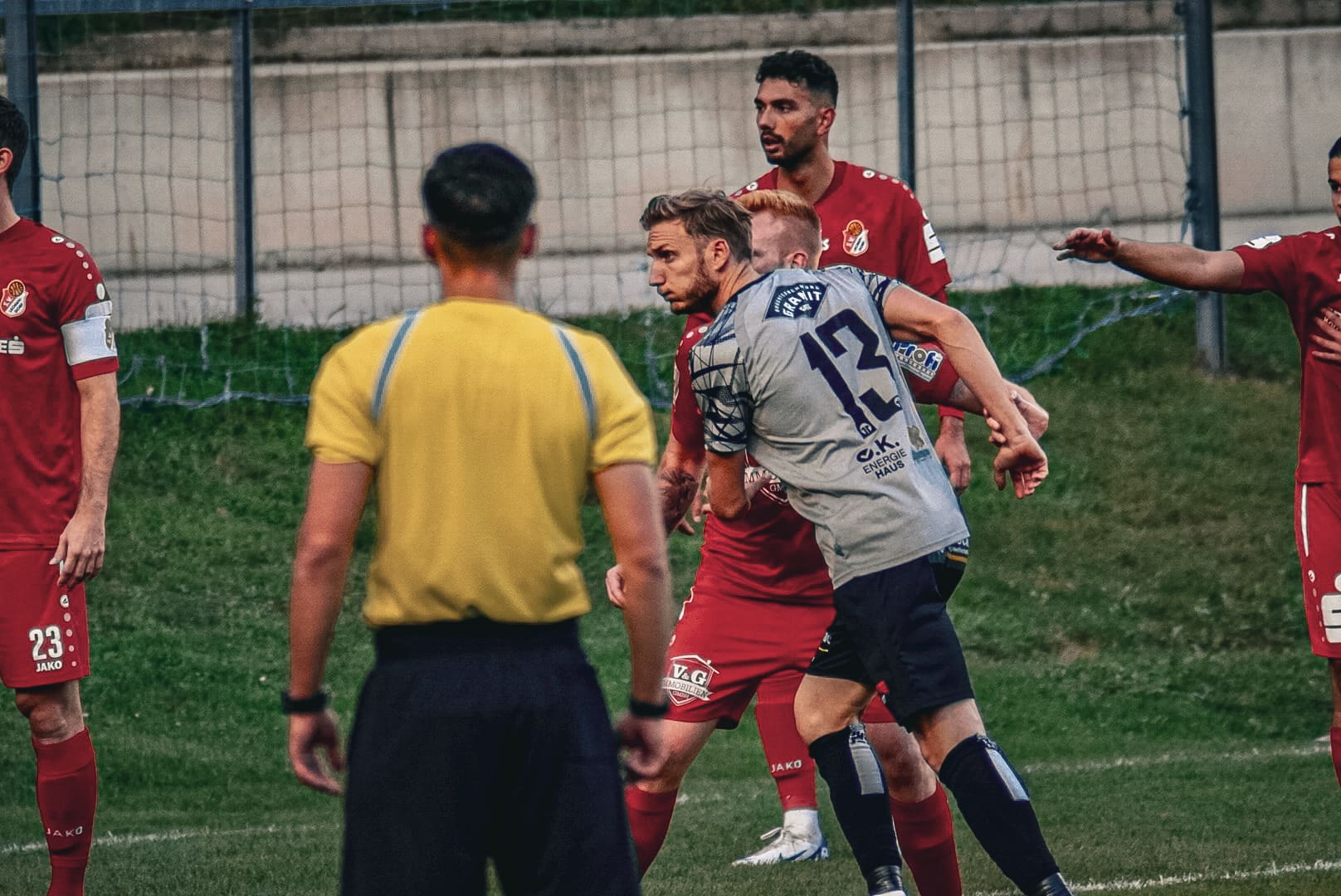 Fußballspiel: Spieler in roten und grauen Trikots im intensiven Zweikampf, Schiedsrichter beobachtet die Szene.