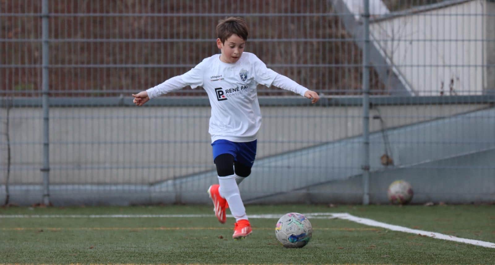 Kind spielt Fußball auf dem Sportplatz, beim Dribbeln mit blauer Hose und weißem Trikot, vor Tor und Ball.