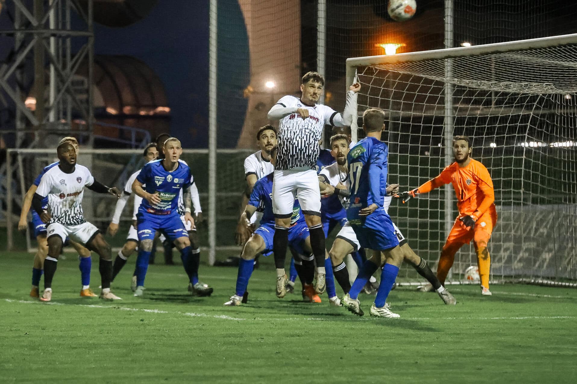 Fußballspiel bei Nacht: Spieler kämpfen im Strafraum um den Ball, Torwart in Aktion, Stadionatmosphäre.