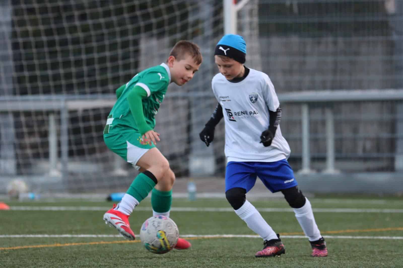 Kinder spielen Fußball auf dem Spielfeld, im grünen und weißen Trikot, konzentriert auf den Ball im Zweikampf.