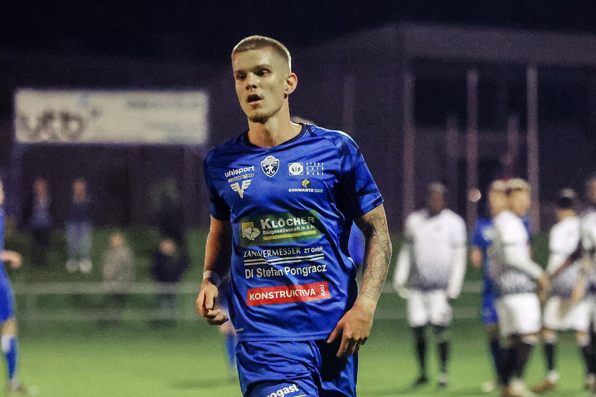 Fußballspieler im blauen Trikot während eines Nachtsportwettbewerbs auf dem Spielfeld, konzentriert und engagiert.