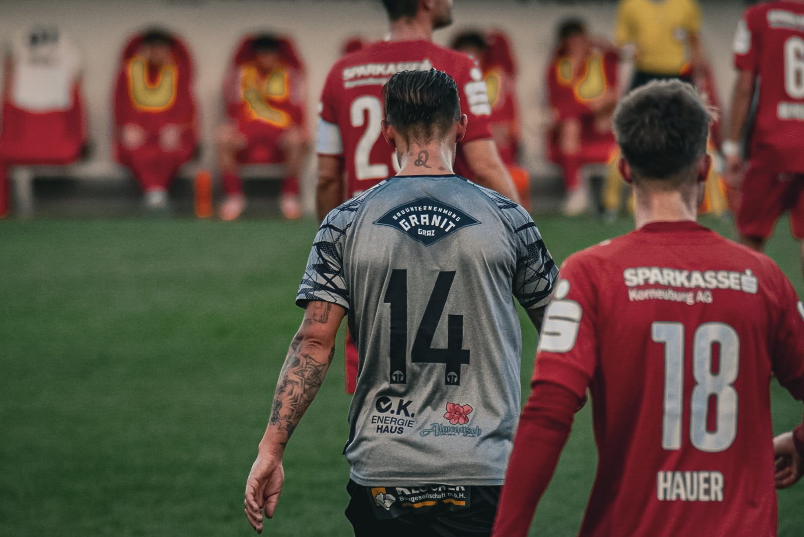 Fußballspieler in roten und grauen Trikots auf dem Spielfeld während eines Spiels, Bank im Hintergrund.