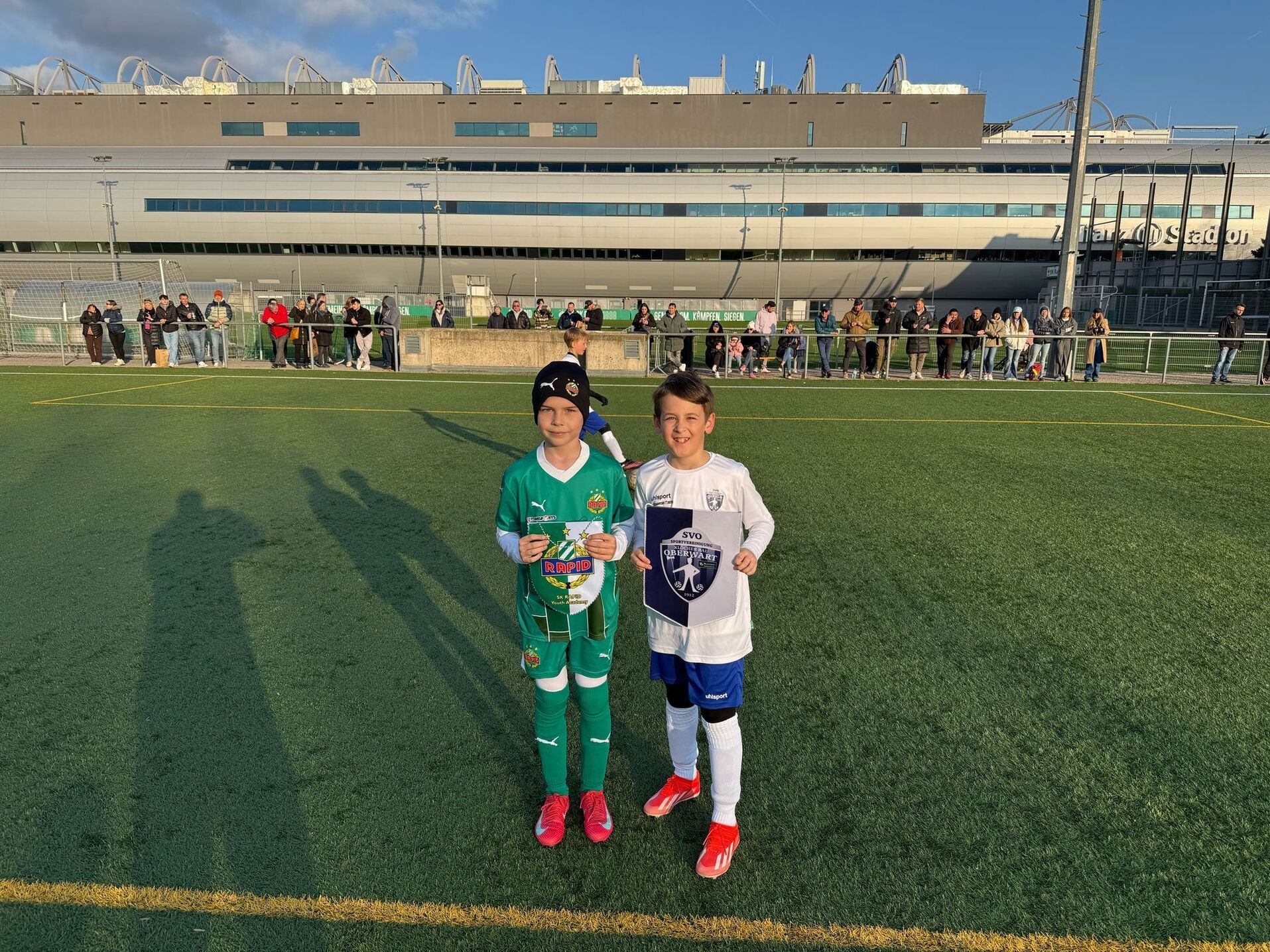 Zwei junge Fußballspieler posieren auf einem Sportplatz, umgeben von Zuschauern und einem Stadion im Hintergrund.