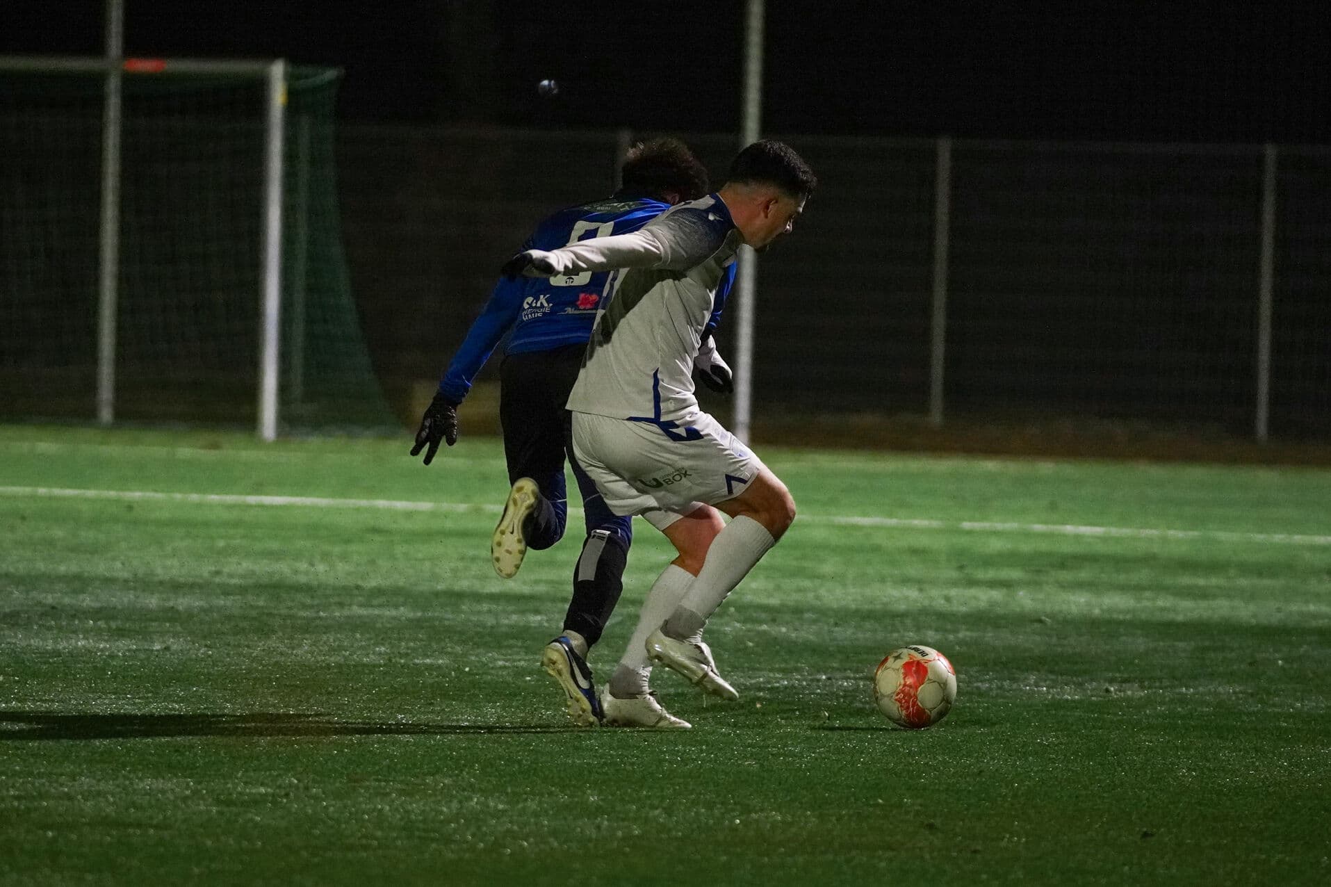 Zwei Fußballspieler kämpfen um den Ball auf einem beleuchteten Spielfeld bei Nacht.