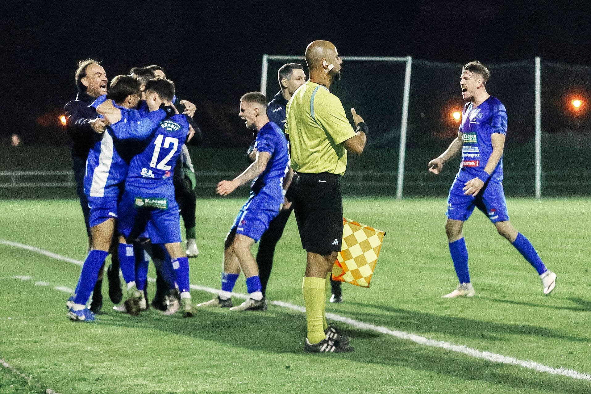 Fußballspieler feiern Torerfolg bei Nachtspiel, Schiedsrichterassistent schaut zu. Spannung und Jubel auf dem Spielfeld.