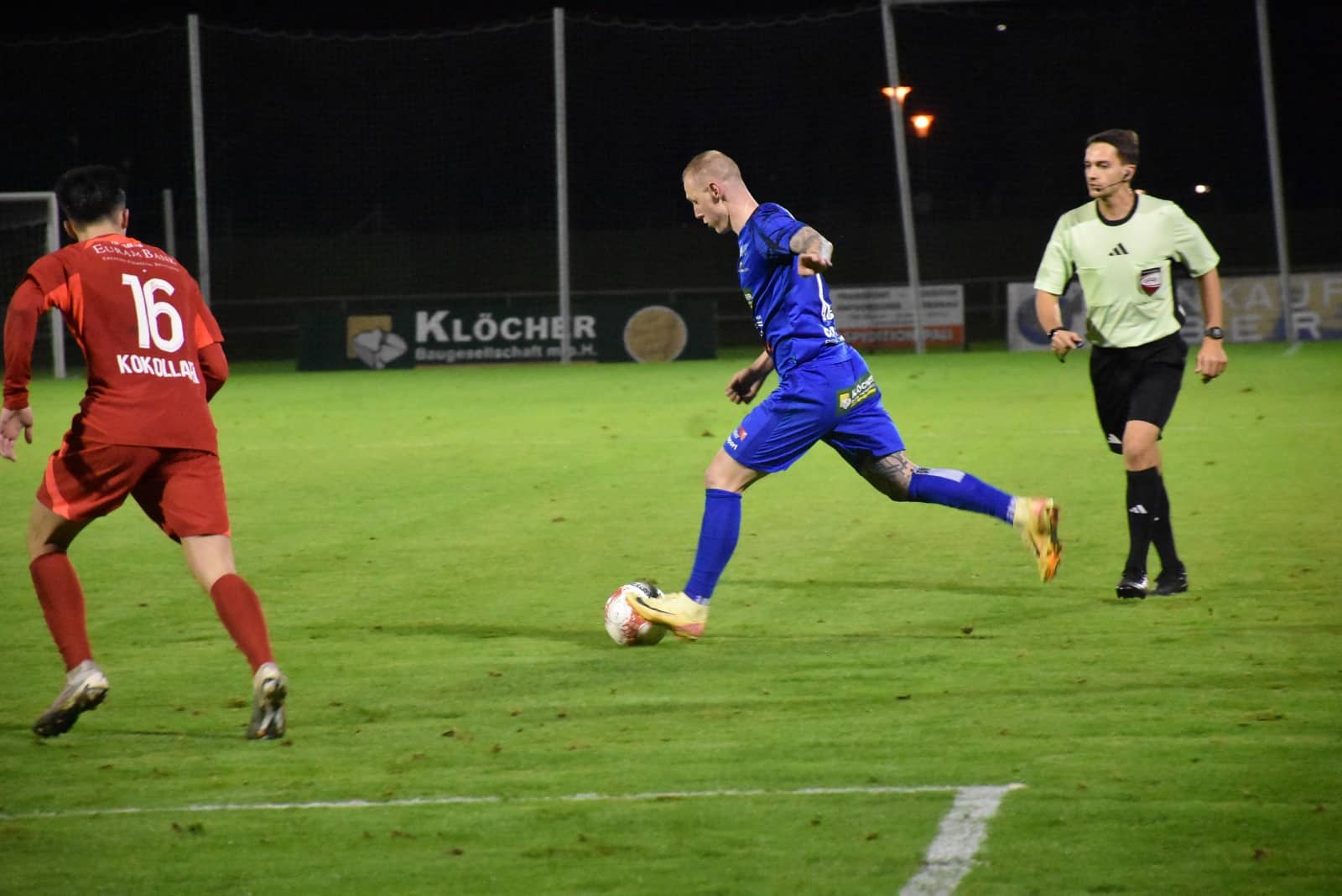 Fußballspiel bei Nacht: Spieler im blauen Trikot dribbelt vor Schiedsrichter und Gegenspieler in rotem Trikot.