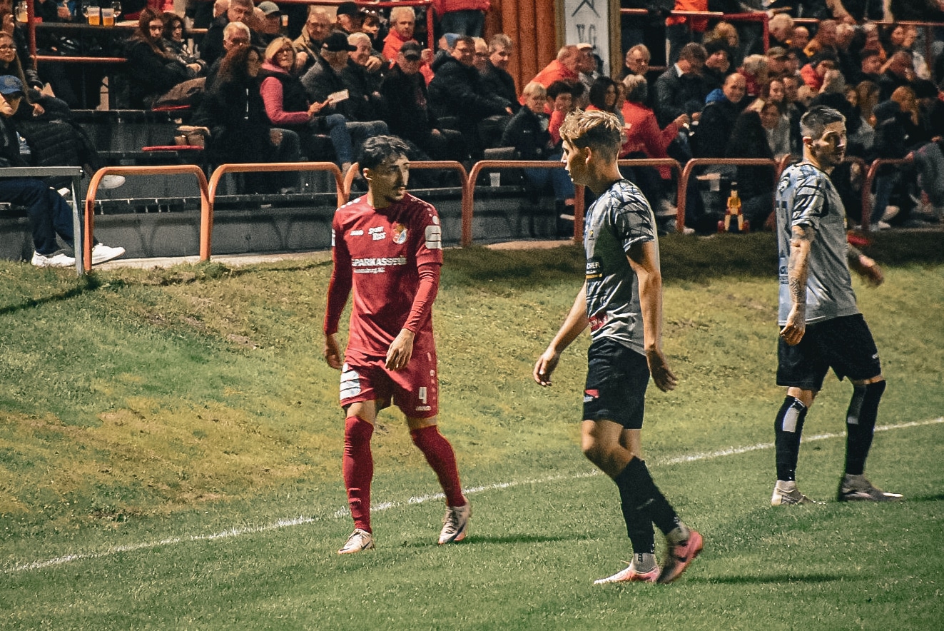 Fußballspieler im roten Trikot während eines Spiels, Zuschauer auf Tribüne im Hintergrund.