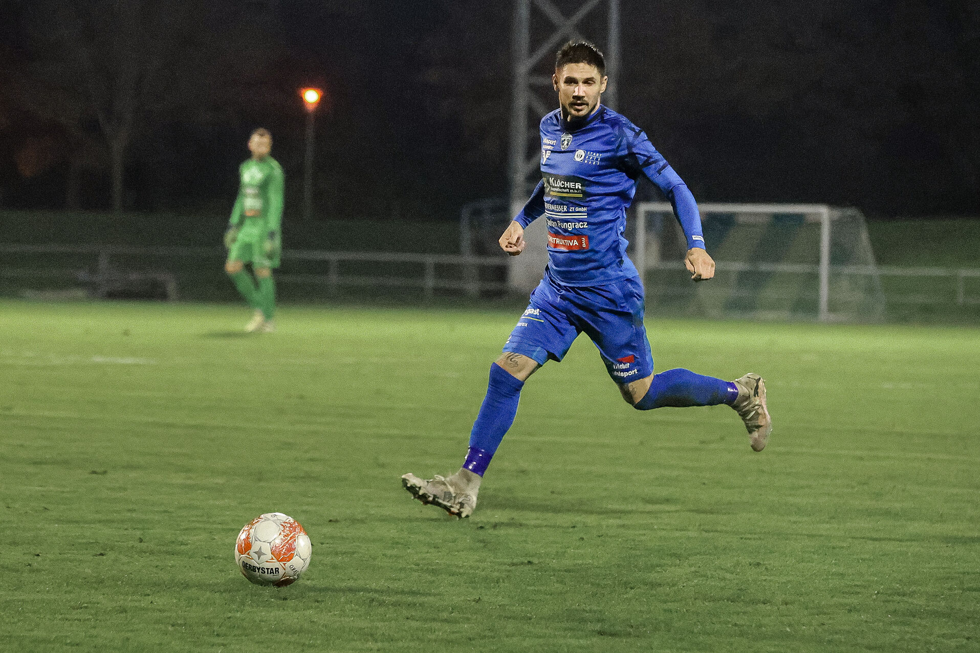 Fußballspieler in blauem Trikot dribbelt den Ball auf dem Spielfeld bei Nacht, Torwart im Hintergrund.