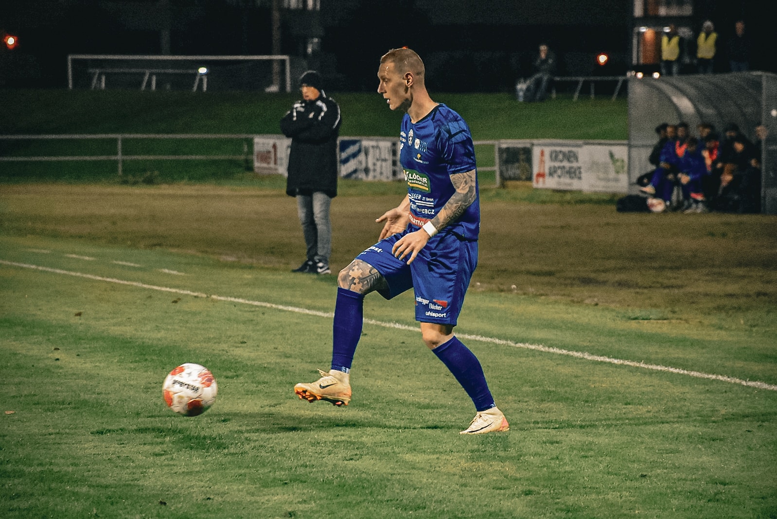 Fußballspieler in blauem Trikot dribbelt auf dem Spielfeld bei Nachtspiel, Trainerteam im Hintergrund.