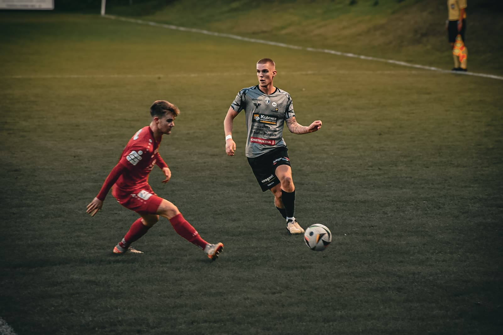 Zwei Fußballspieler im Zweikampf auf dem Spielfeld, einer in Rot, der andere in Grau, mit einem Fußball zwischen ihnen.