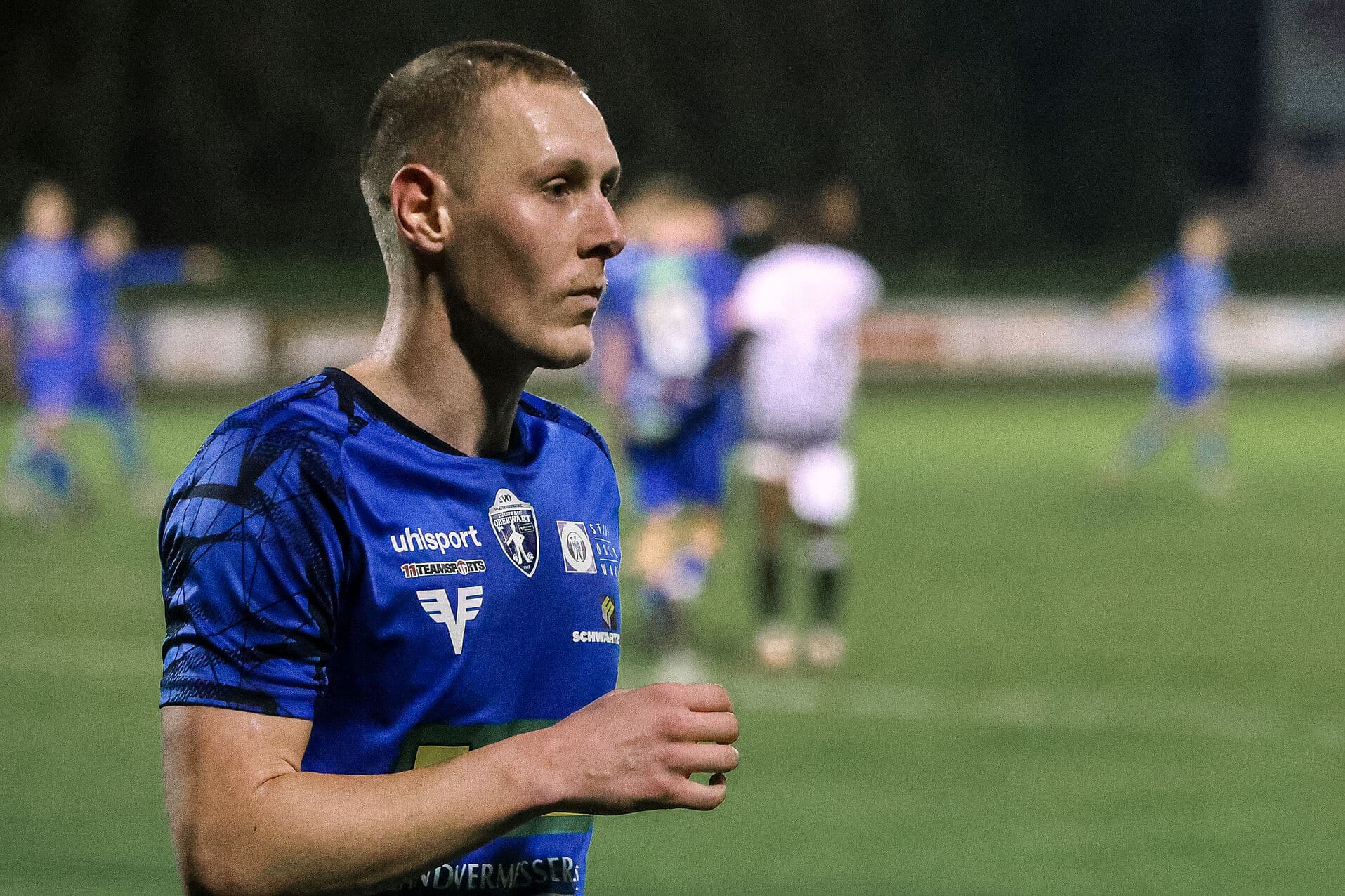 Ein Fußballspieler im blauen Trikot auf dem Spielfeld konzentriert während eines Spiels, mit unscharfen Spielern im Hintergrund.
