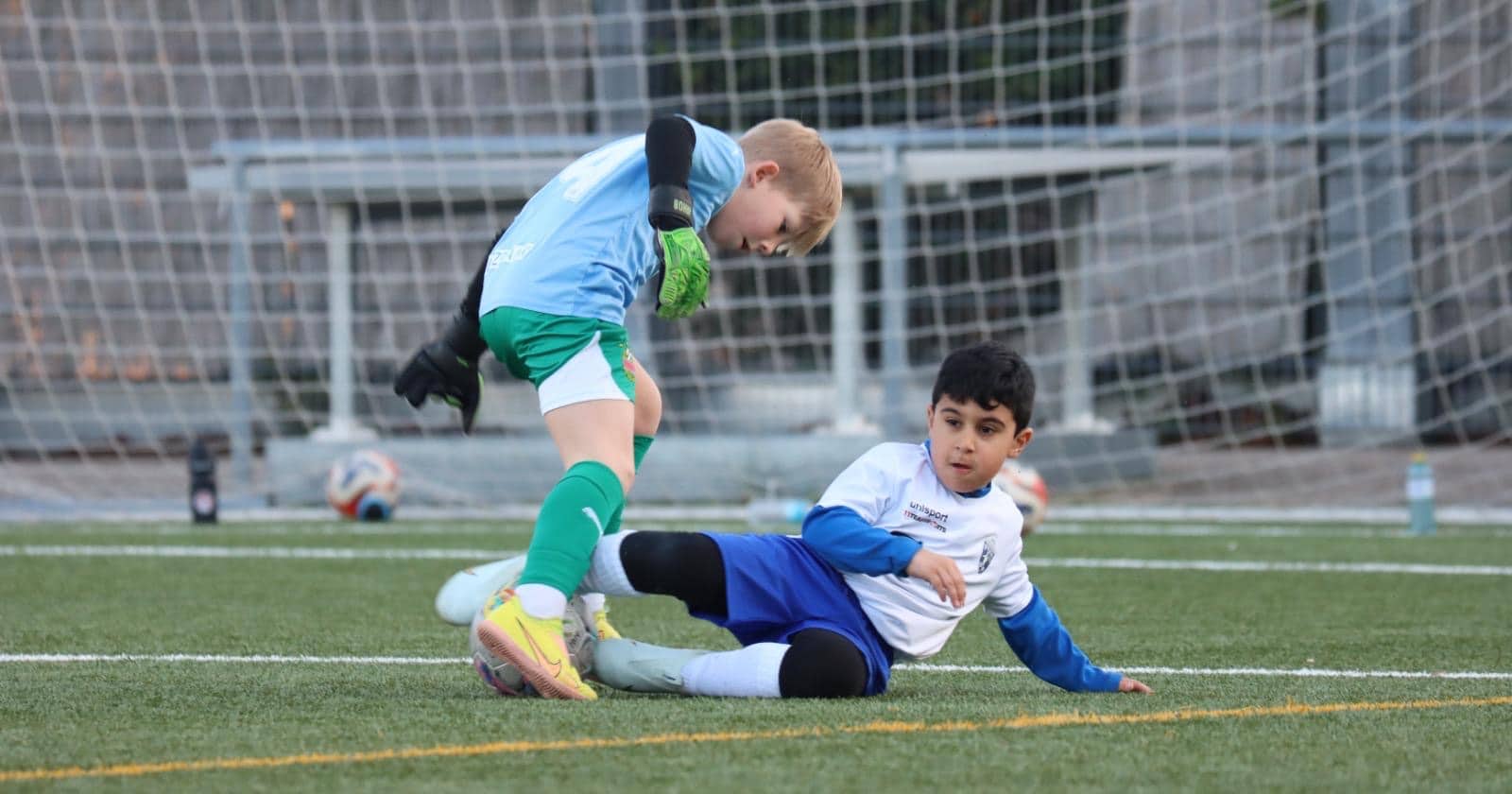 Zwei Kinder spielen Fußball, eins im Torwarttrikot in Aktion vor dem Tor auf einem Sportplatz.