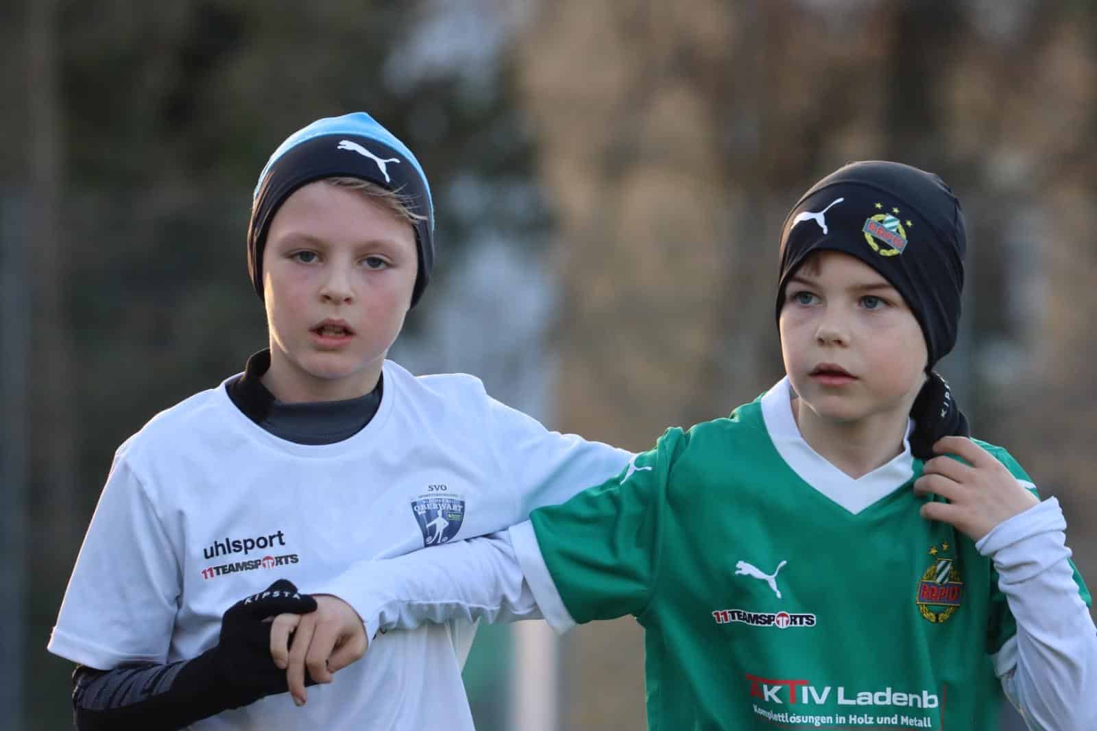 Zwei junge Fußballspieler im Einsatz, gekleidet in weiße und grüne Trikots mit schwarzen Mützen, konzentriert auf das Spiel.