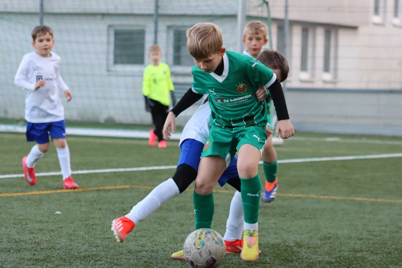 Kinder spielen Fußball auf einem Platz, im Fokus ist ein Zweikampf um den Ball zwischen zwei Spielern in Grün und Weiß.