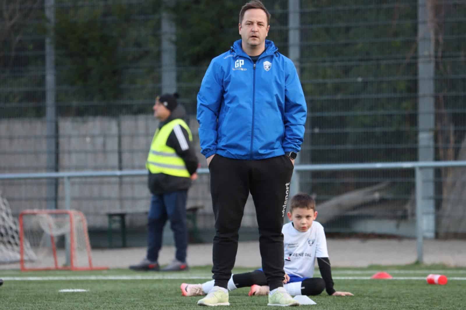 Fußballtrainer in blauer Jacke beobachtet Jugendtraining auf dem Spielfeld, während ein Junge auf dem Boden sitzt.