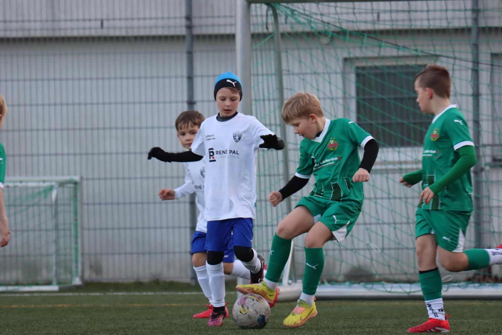 Kinder spielen Fußballspiel im Freien, in weißen und grünen Trikots auf einem Rasenfeld, nahe dem Tor.
