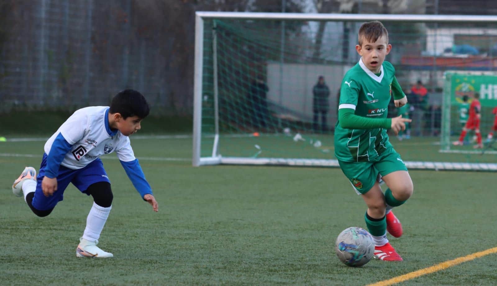 Junger Fußballer in grünem Trikot dribbelt den Ball, während Gegenspieler im weißen Trikot versucht, ihn abzufangen.
