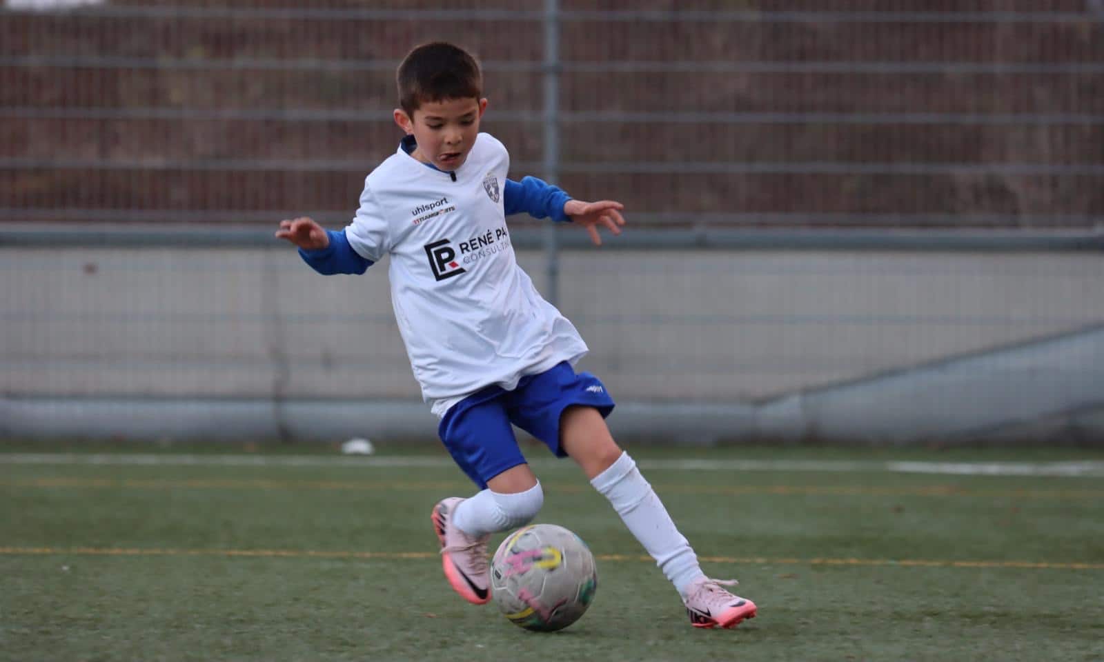 Junger Fußballer in Aktion auf dem Spielfeld, trägt ein weißes Trikot und blaue Shorts, bereit für einen Pass.