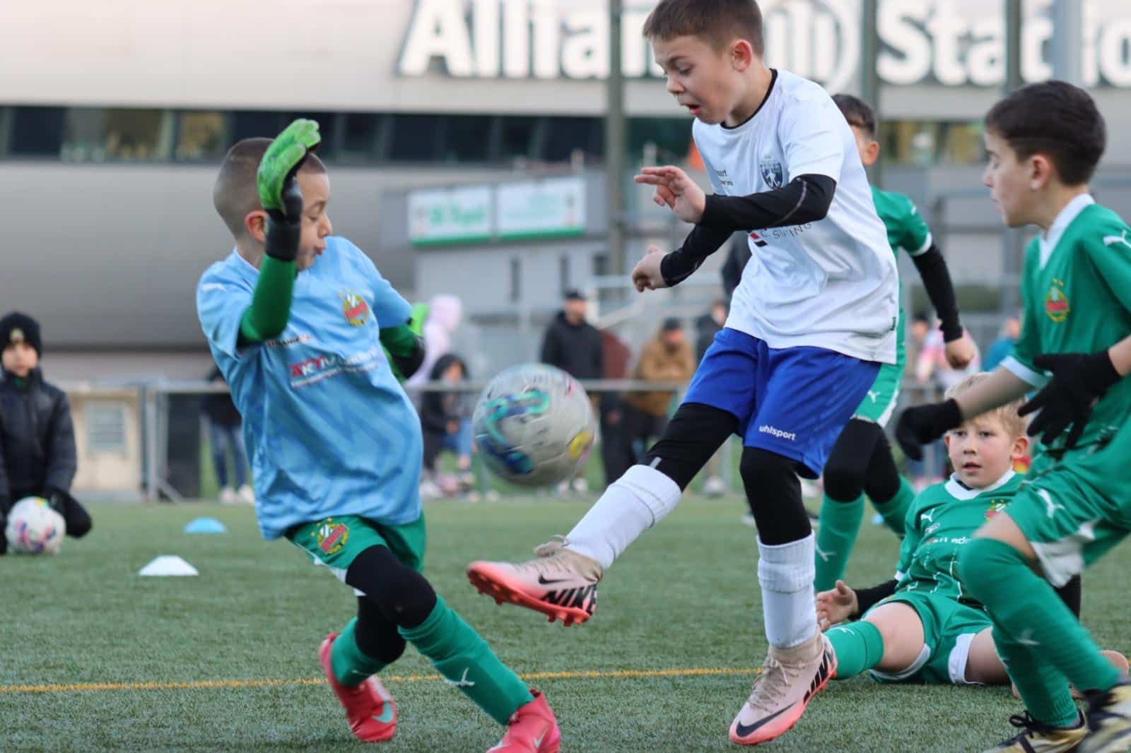 Kinder beim Fußballspielen auf einem Sportplatz vor einem Allianz-Gebäude, voller Einsatz beim Kampf um den Ball.