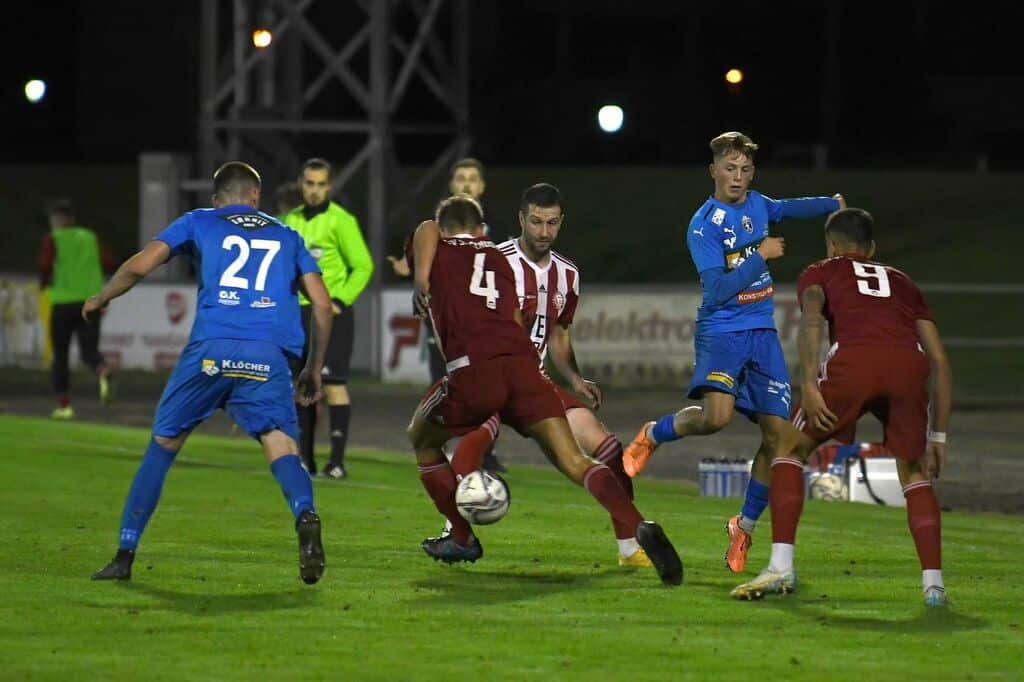 Fußballspiel bei Nacht: Spieler in Blau und Rot kämpfen energisch um den Ball auf einem beleuchteten Spielfeld.