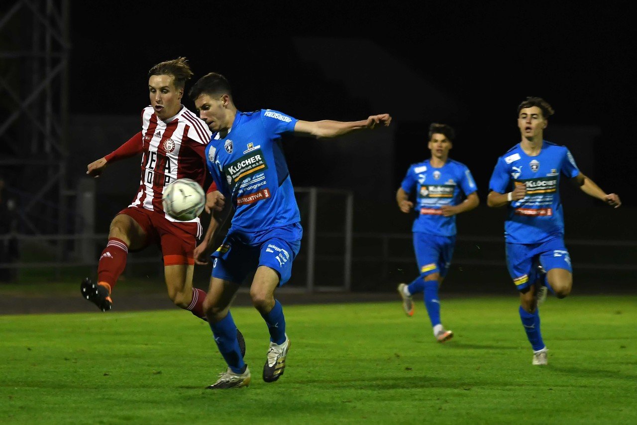Fußballspiel bei Nacht: Spieler im Zweikampf um den Ball, einer in Rot, einer in Blau, auf grünem Rasen.