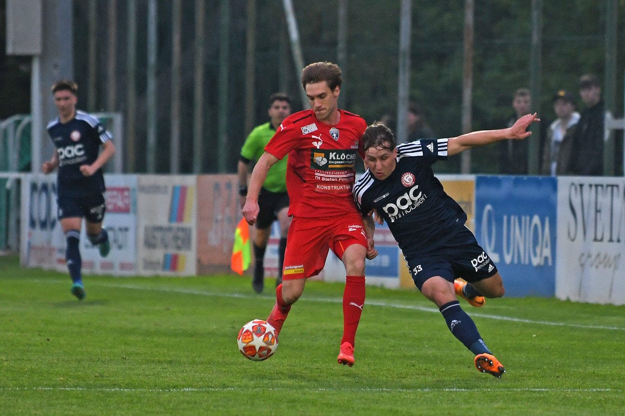 Zwei Fußballspieler kämpfen um den Ball auf einem Spielfeld, während ein Schiedsrichter im Hintergrund beobachtet.