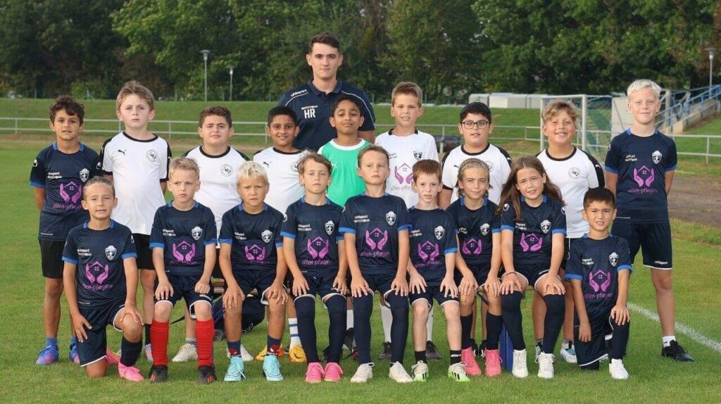 Jugendfußballteam posiert auf dem Spielfeld, 15 Jungen in blauen Trikots und weißen Trikots, mit Trainer.