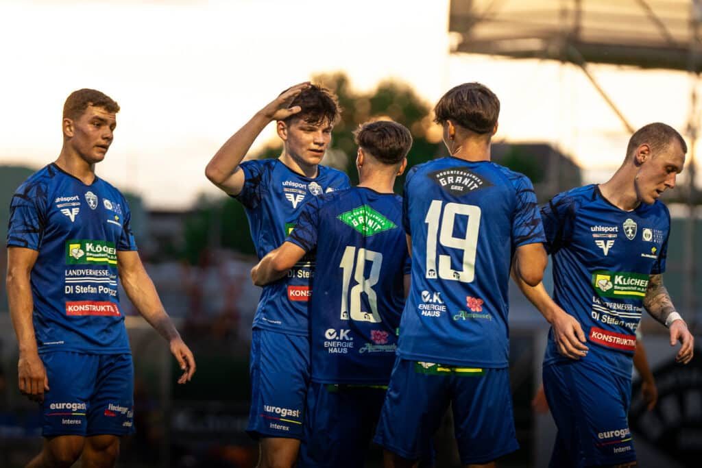 Fußballspieler im blauen Trikot feiern auf dem Spielfeld bei Sonnenuntergang, Teamjubel, Sportereignis.
