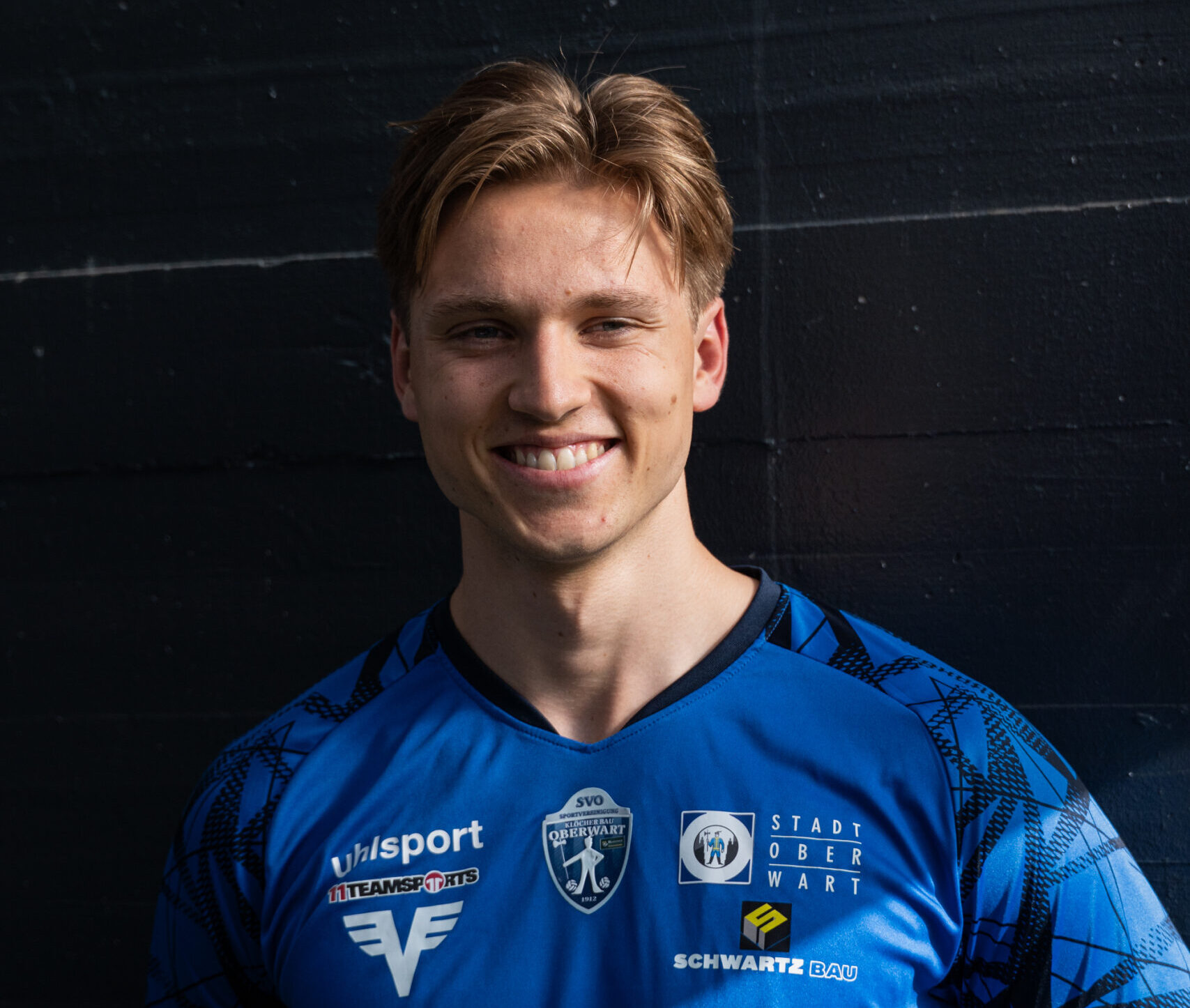 Ein Spieler im blauen Fußballtrikot von SVO Oberwart lächelt in die Kamera vor dunklem Hintergrund.