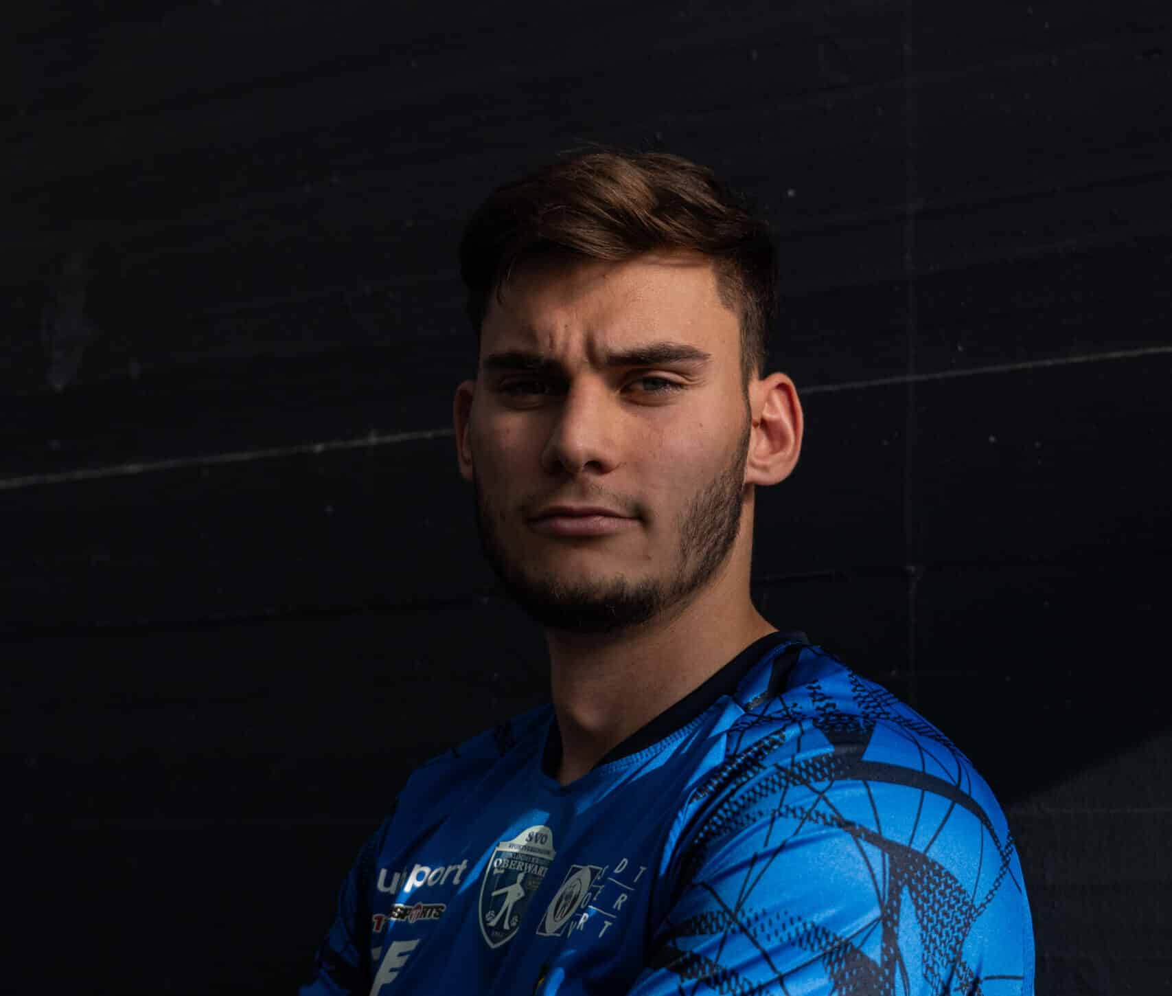 Sportler im blauen Trikot steht mit verschränkten Armen vor dunklem Hintergrund und schaut selbstbewusst zur Kamera.