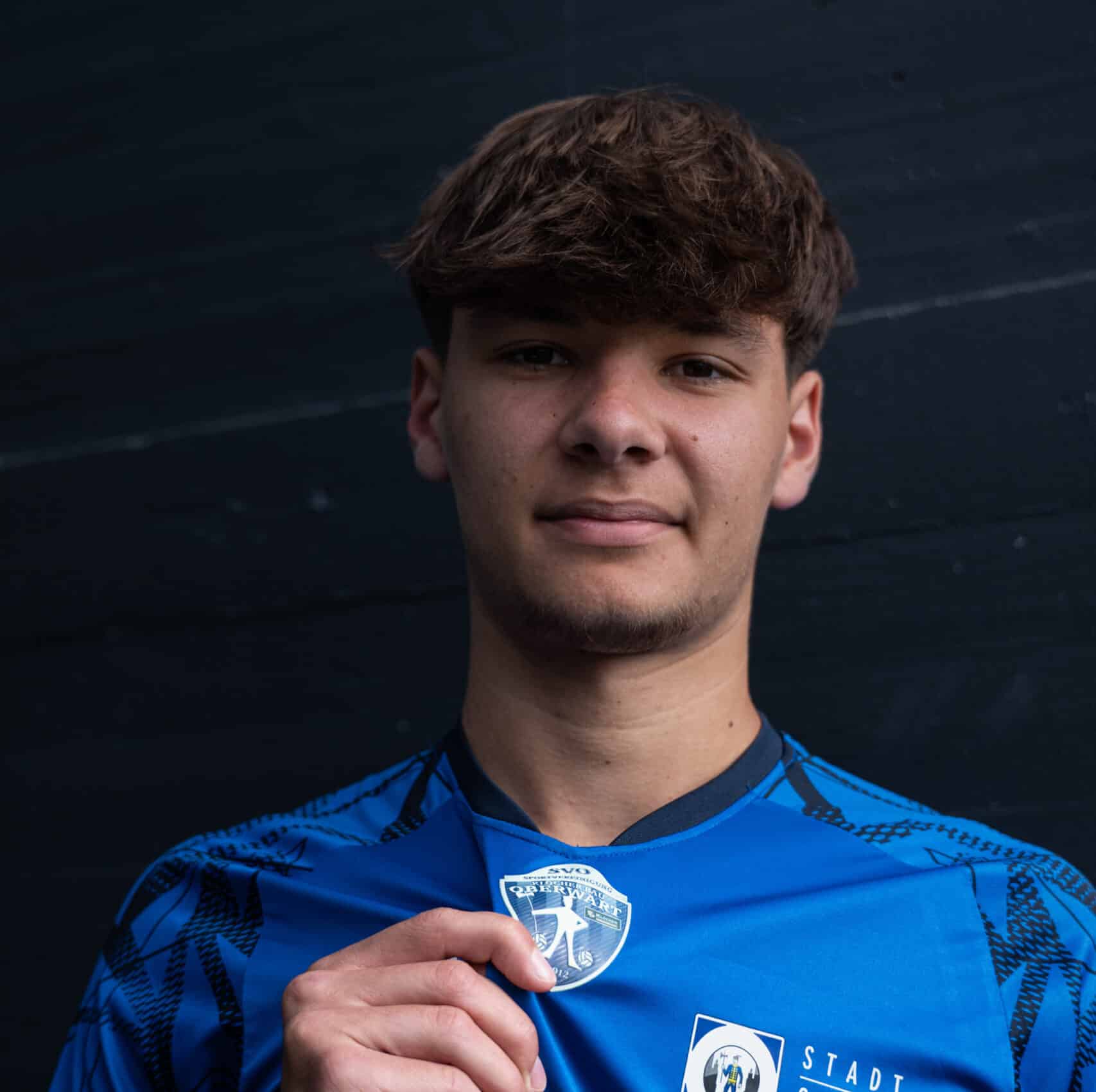 Junger Fußballspieler im blauen Trikot mit Vereinswappen, stolz vor dunklem Hintergrund, zeigt Teamstolz.