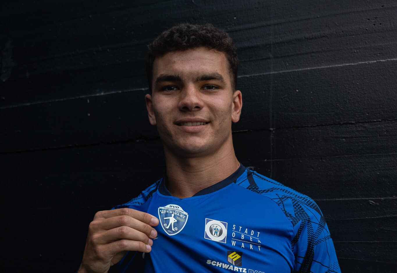 Fußballspieler im blauen Trikot zeigt stolz das Logo seines Teams vor schwarzem Hintergrund.
