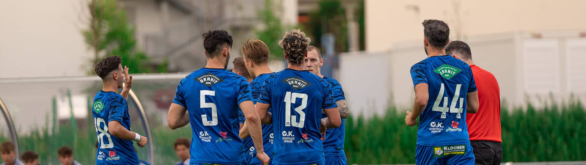 Fußballspieler in blauen Trikots feiern ein Tor auf dem Spielfeld während eines Spiels im Freien.