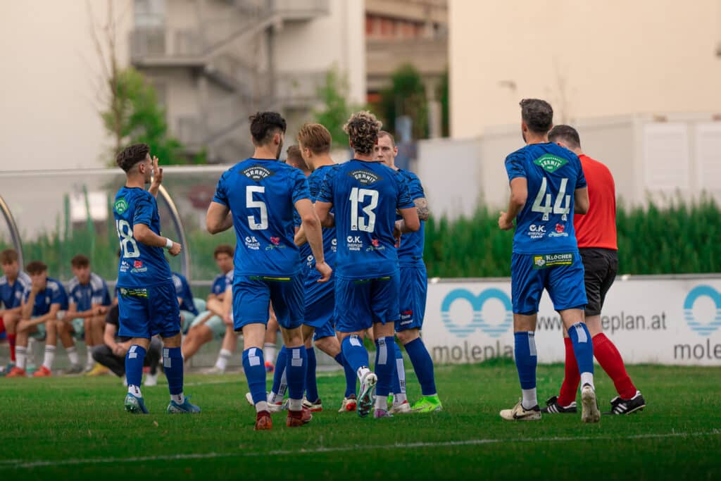 Fußballspieler im blauen Trikot feiern ein Tor auf dem Fußballfeld, mit Mannschaftskollegen im Hintergrund auf der Bank.