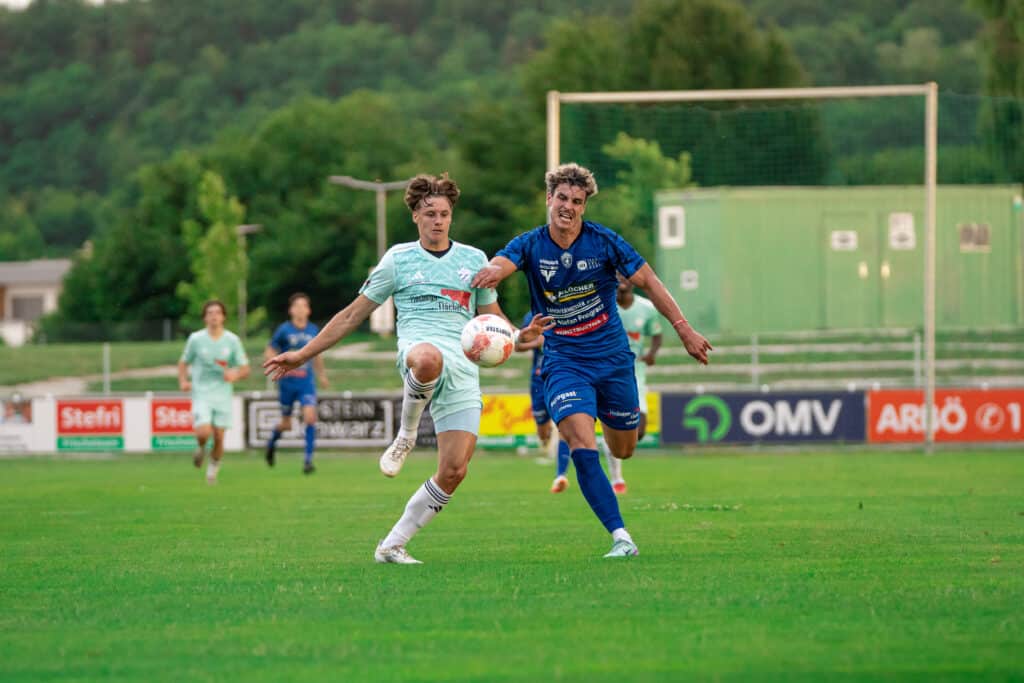 Zwei Fußballspieler im Zweikampf um den Ball auf einem Feld, umgeben von grüner Natur und Stadionatmosphäre.