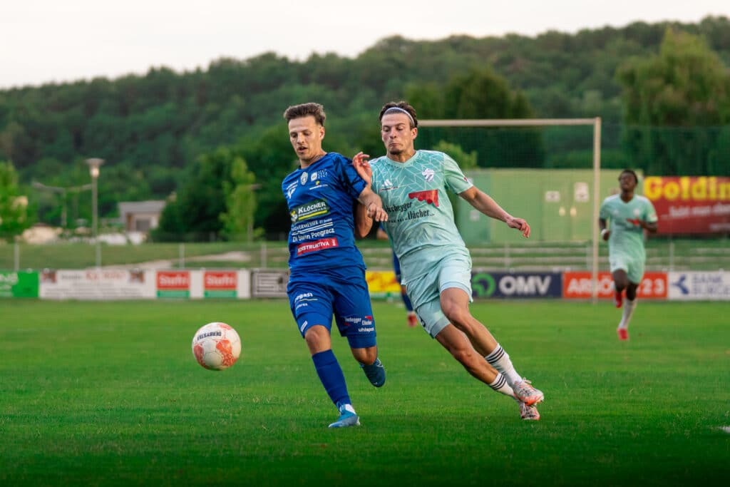 Zwei Fußballspieler kämpfen um den Ball auf dem Spielfeld, während ein dritter Spieler im Hintergrund rennt.