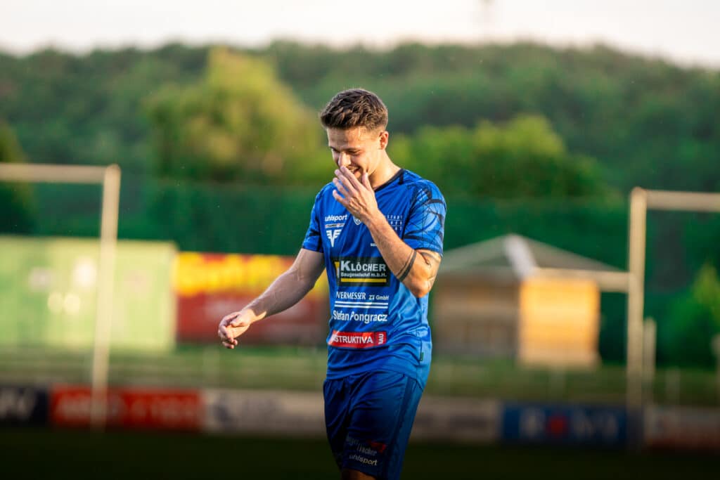 Fußballspieler im blauen Trikot auf dem Spielfeld bei Sonnenuntergang, lächelnd und nachdenklich.