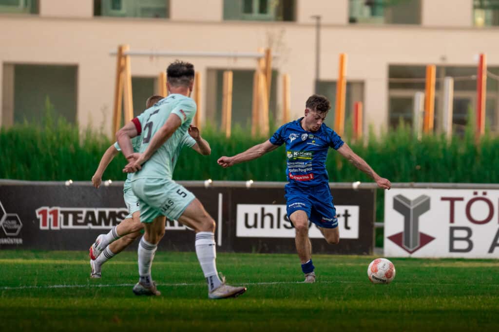 Fußballspiel in Aktion: Spieler in blauem Trikot dribbelt gegen Verteidiger in hellem Trikot auf grünem Rasen.
