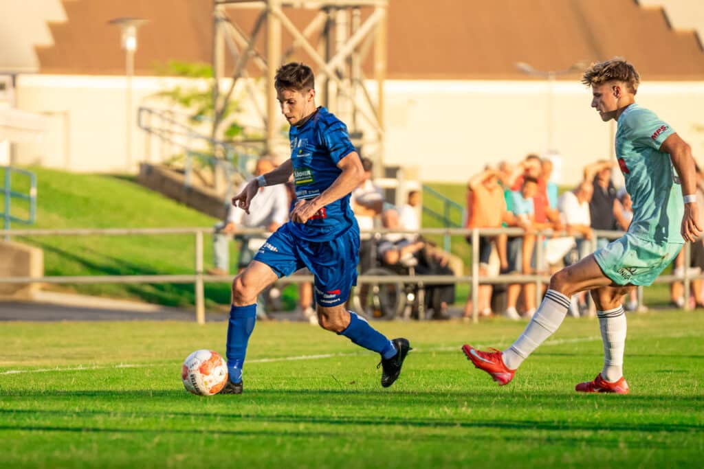 Fußballspiel bei Sonnenschein: Spieler in blau und grün im Zweikampf auf dem Rasen, Tribüne im Hintergrund.