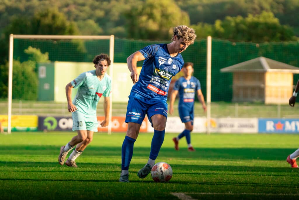 Fußballspieler im blauen Trikot kontrolliert den Ball, während Gegner in mintgrüner Ausrüstung zuschaut, auf grünem Spielfeld.
