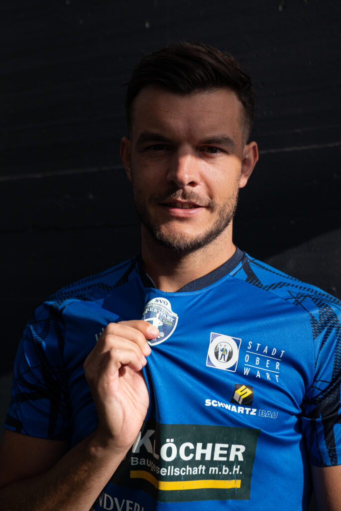 Fußballspieler im blauen Trikot hält stolz sein Vereinslogo in der Hand, symbolisiert Teamgeist und Leidenschaft.